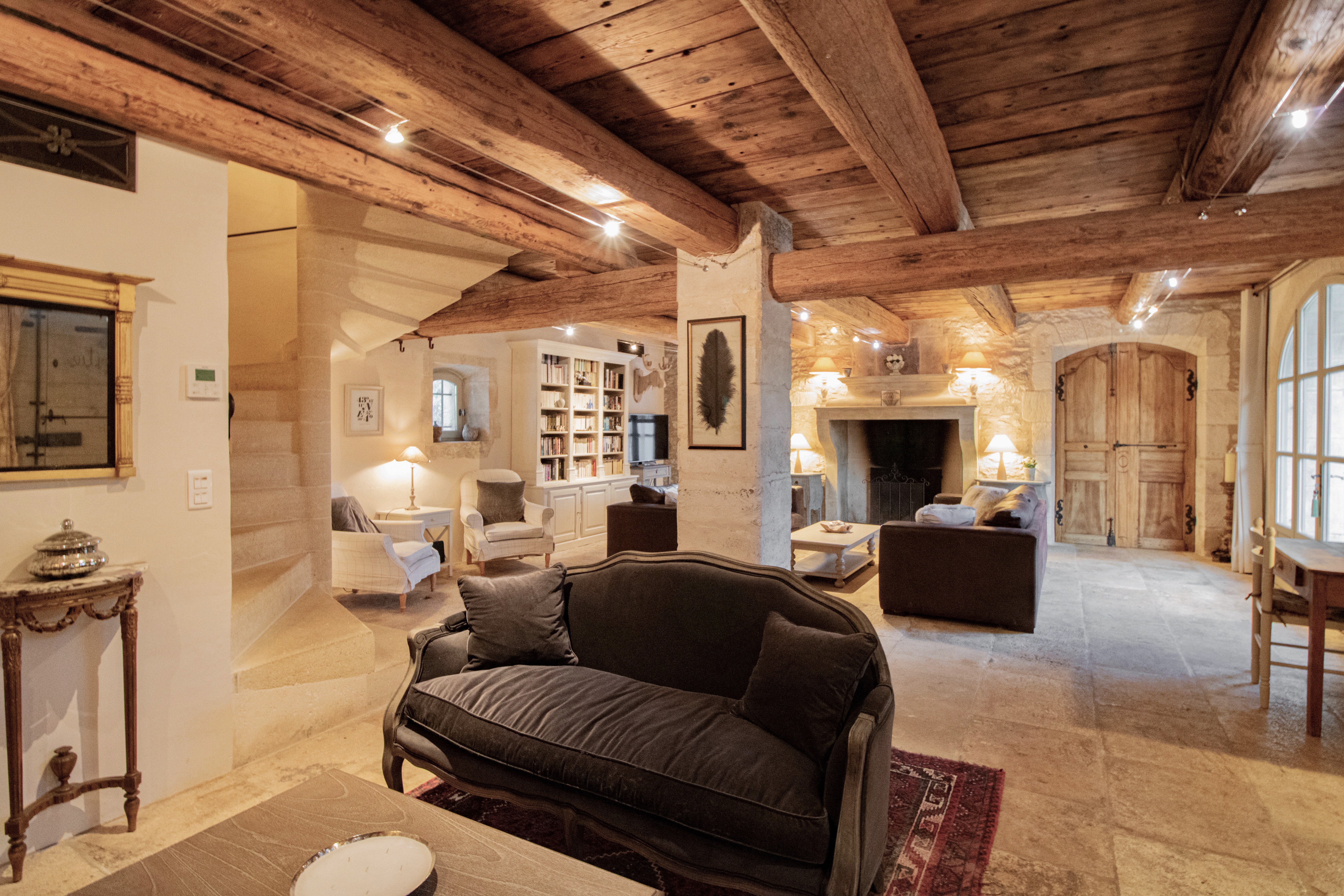 Spacious rustic living room with exposed wooden beams, stone walls, and a fireplace. includes a dark leather sofa, bookshelves, and elegant decor. Warm lighting enhances the cozy ambiance.